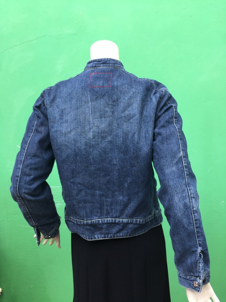 Peut inclure: Une veste en jean bleu avec un petit patch carr&eacute; au dos. La veste a des manches longues et une fermeture &agrave; boutons.