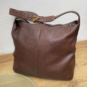 May include: A brown leather shoulder bag with a single strap and a gold-colored V-shaped buckle. The bag has a large, slouchy design and a zipper closure. The leather appears to be textured.