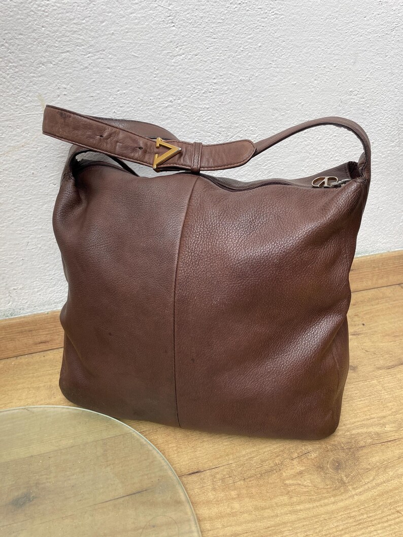 May include: A brown leather shoulder bag with a single strap and a gold-colored V-shaped buckle. The bag has a large, slouchy design and a zipper closure. The leather appears to be textured.