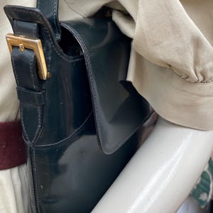 Puede incluir: Un bolso de hombro de color verde oscuro con un acabado liso y brillante y cierre de solapa. El bolso cuenta con una correa de cuero vertical con una hebilla de metal dorado para ajustar. El dise&ntilde;o incluye una correa larga para el hombro y un cuerpo rectangular estructurado.
