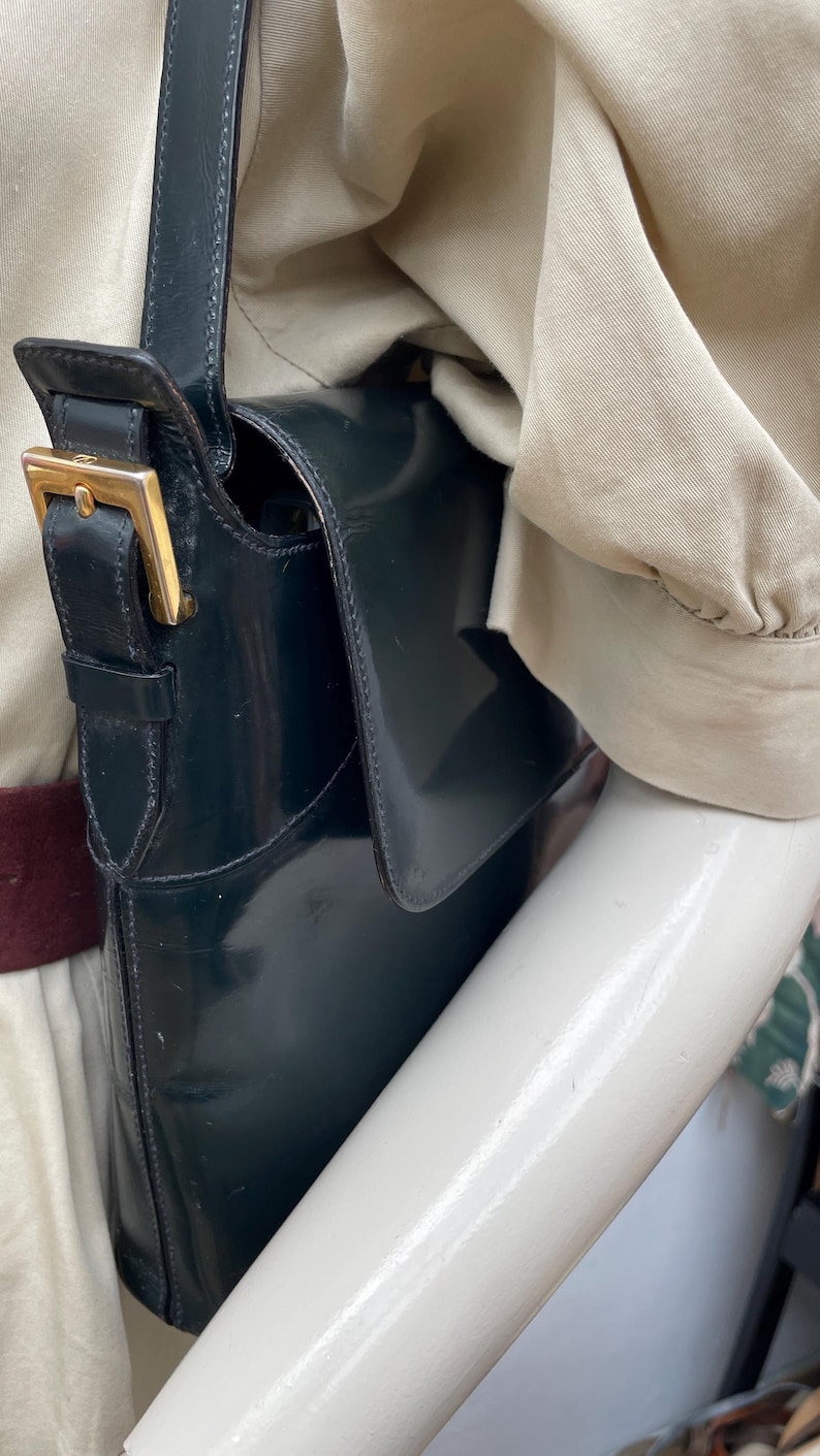 Puede incluir: Un bolso de hombro de color verde oscuro con un acabado liso y brillante y cierre de solapa. El bolso cuenta con una correa de cuero vertical con una hebilla de metal dorado para ajustar. El dise&ntilde;o incluye una correa larga para el hombro y un cuerpo rectangular estructurado.