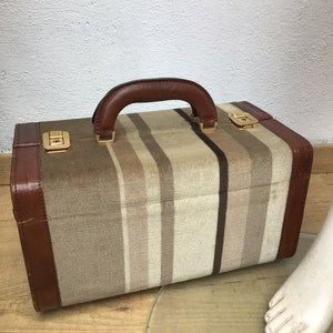 Op de afbeelding: Een vintage rechthoekige koffer met een bruine leren handgreep en rand. De behuizing van de koffer heeft verticale strepen in tinten beige, bruin en crème. Goudkleurige sluitingen zekeren het deksel.