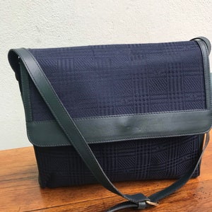 May include: A navy blue crossbody bag with a plaid pattern. The bag features a flap closure and a dark green leather strap and trim. The bag is sitting on a wooden surface.