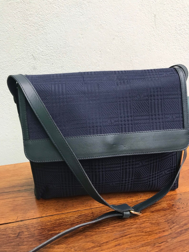 May include: A navy blue crossbody bag with a plaid pattern. The bag features a flap closure and a dark green leather strap and trim. The bag is sitting on a wooden surface.