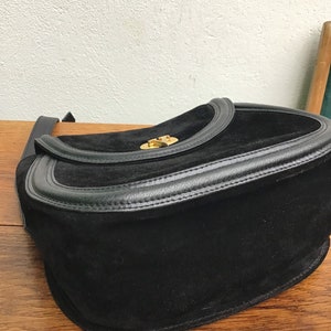 May include: Black suede shoulder bag with a leather trim and a gold-tone clasp. The bag has a flap closure and a long strap. The bag is sitting on a wooden surface.