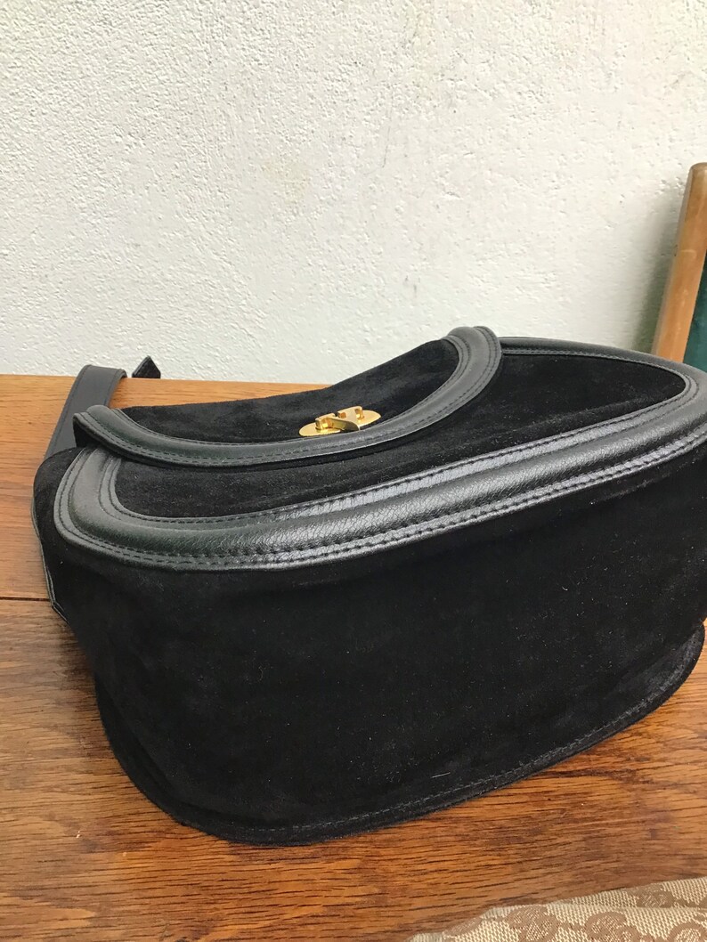 May include: Black suede shoulder bag with a leather trim and a gold-tone clasp. The bag has a flap closure and a long strap. The bag is sitting on a wooden surface.
