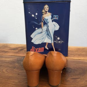 May include: A pair of brown leather heeled mules with a square toe, set in front of a vintage blue Dixan tin featuring a woman in a white dress. The shoes have a block heel.