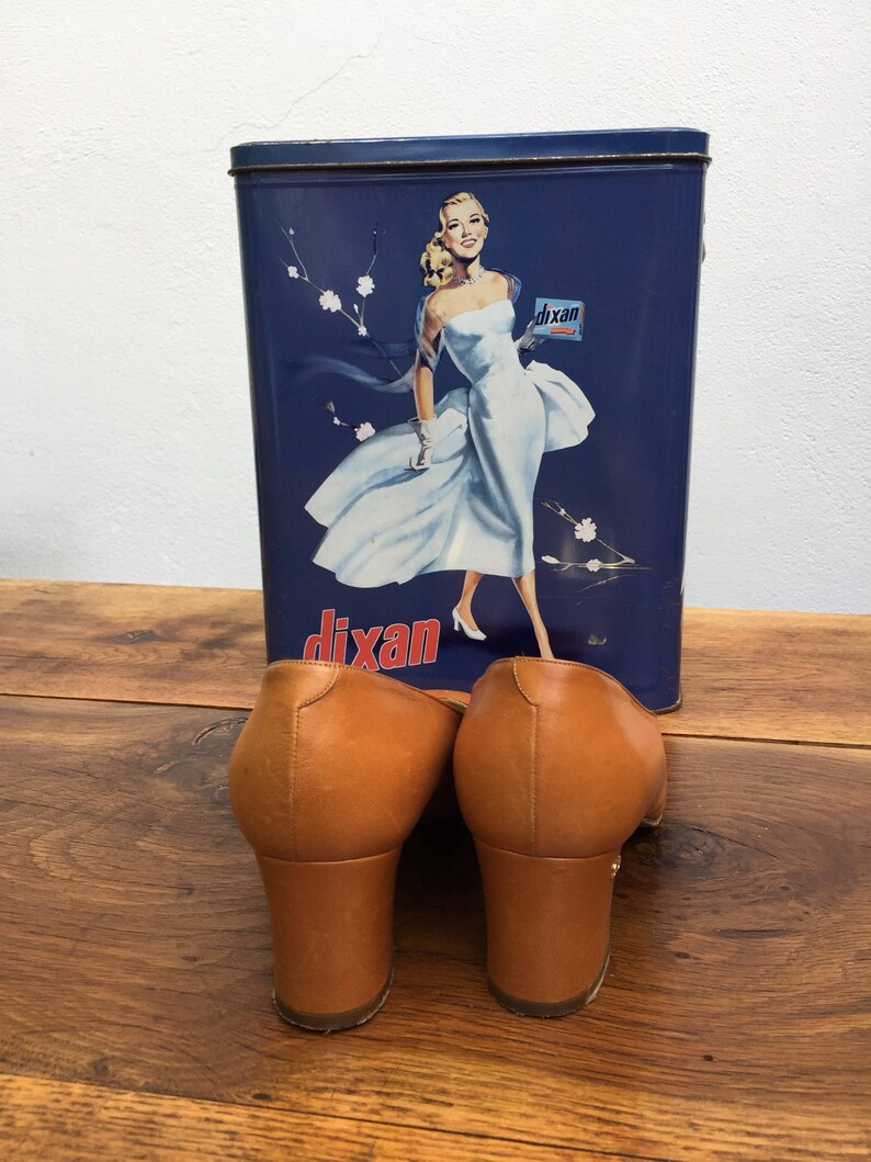 May include: A pair of brown leather heeled mules with a square toe, set in front of a vintage blue Dixan tin featuring a woman in a white dress. The shoes have a block heel.