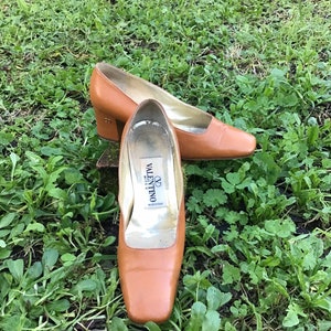 May include: A pair of brown leather pumps with a square toe and a low heel. The shoes are set on a bed of green grass. The inside of one shoe has a label that reads "Valentino Miss V".