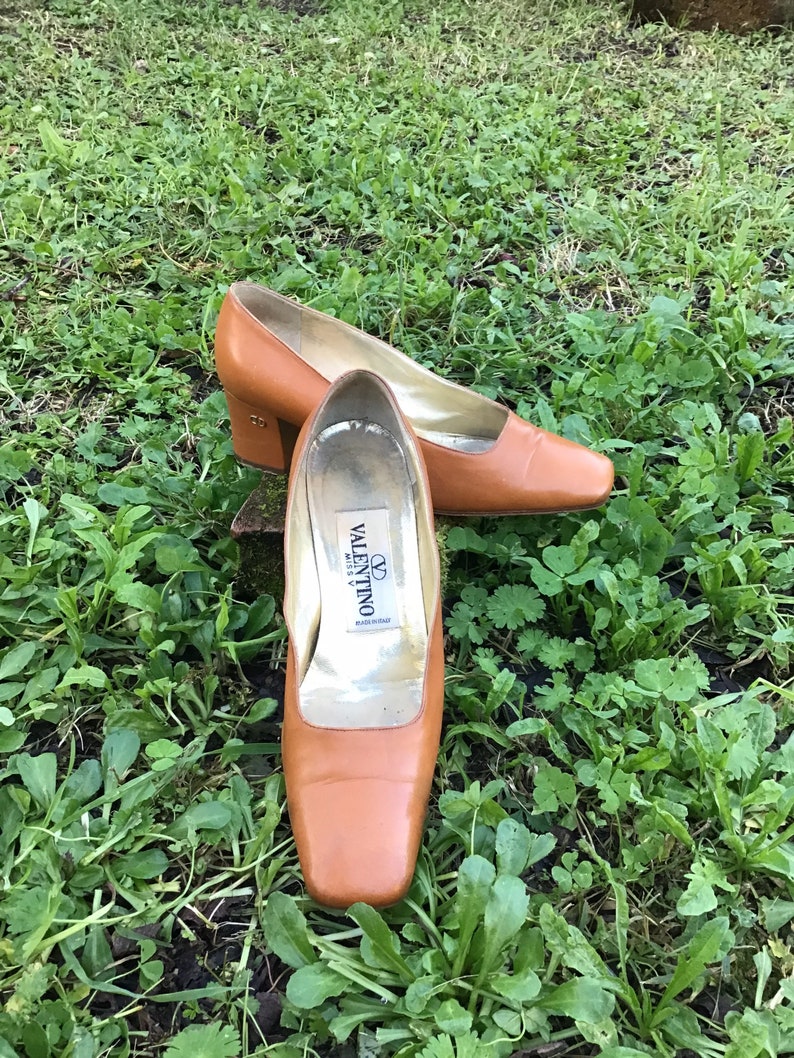 May include: A pair of brown leather pumps with a square toe and a low heel. The shoes are set on a bed of green grass. The inside of one shoe has a label that reads "Valentino Miss V".