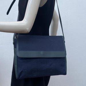 May include: A navy blue shoulder bag with a plaid pattern. The bag has a flap closure and a long, adjustable strap. The bag is displayed on a mannequin wearing a black dress.