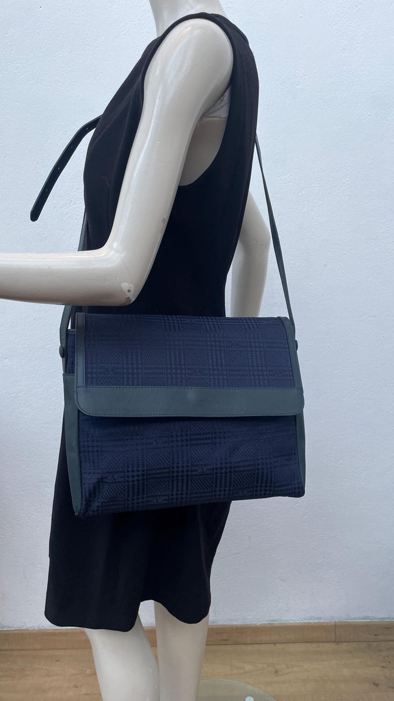 May include: A navy blue shoulder bag with a plaid pattern. The bag has a flap closure and a long, adjustable strap. The bag is displayed on a mannequin wearing a black dress.