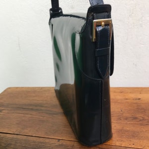 Puede incluir: Un bolso de hombro azul marino oscuro con un acabado brillante de charol. El bolso presenta una silueta rectangular estructurada, un cierre de solapa asegurado con una hebilla de metal dorado y una correa a juego en el lateral.