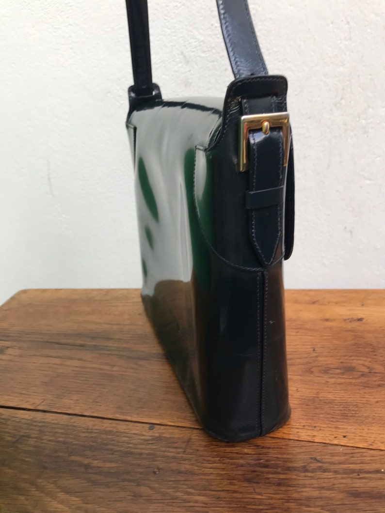 Puede incluir: Un bolso de hombro azul marino oscuro con un acabado brillante de charol. El bolso presenta una silueta rectangular estructurada, un cierre de solapa asegurado con una hebilla de metal dorado y una correa a juego en el lateral.