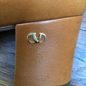 May include: Close-up of a tan leather shoe heel. The heel is a block style with a gold-colored logo. The shoe appears to be vintage or pre-owned, with some wear visible on the sole.