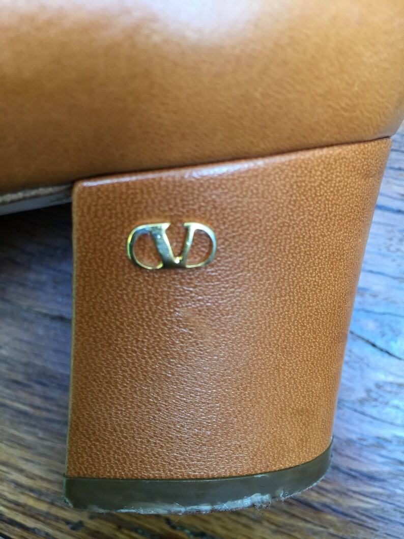 May include: Close-up of a tan leather shoe heel. The heel is a block style with a gold-colored logo. The shoe appears to be vintage or pre-owned, with some wear visible on the sole.