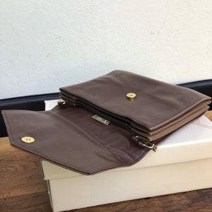 May include: A brown leather clutch with a gold-tone clasp and hardware. The clutch is open, revealing the interior and a small gold logo. The bag is resting on a white box, with a wooden surface below.