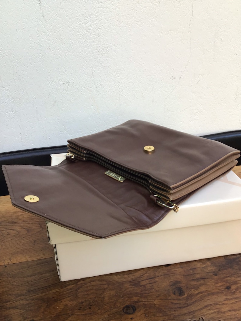 May include: A brown leather clutch with a gold-tone clasp and hardware. The clutch is open, revealing the interior and a small gold logo. The bag is resting on a white box, with a wooden surface below.