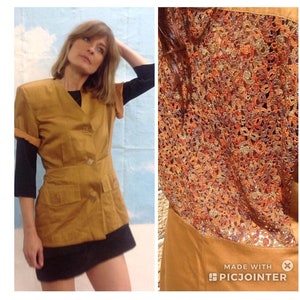 May include: Mustard-coloured short-sleeved jacket with a black undershirt and a black skirt. The jacket has a V-neck, two pockets, and a button closure. The back of the jacket features a detailed, colourful, open-weave design.