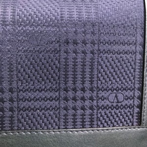 May include: Close-up of a dark blue handbag with a woven, textured pattern. The bag features a geometric design and a dark gray leather trim along the bottom. The strap is visible at the top.