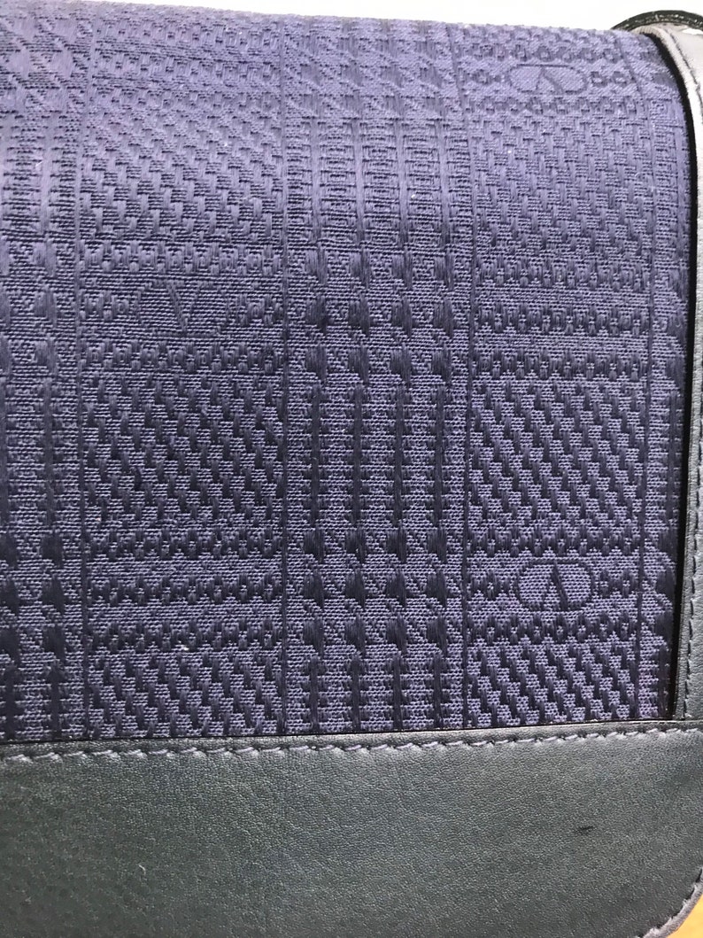 May include: Close-up of a dark blue handbag with a woven, textured pattern. The bag features a geometric design and a dark gray leather trim along the bottom. The strap is visible at the top.