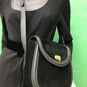 May include: Black suede crossbody bag with a rounded flap and gold-tone clasp. The bag features a black leather trim and a long, adjustable strap. The bag is displayed on a mannequin wearing a black dress.