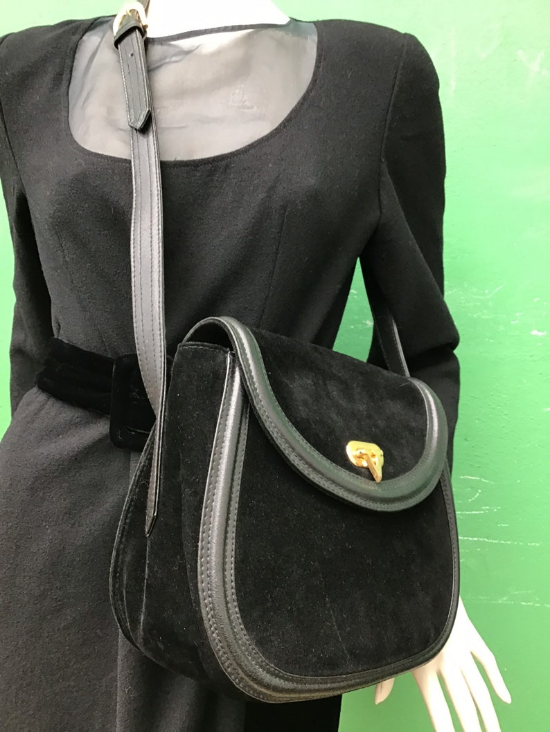 May include: Black suede crossbody bag with a rounded flap and gold-tone clasp. The bag features a black leather trim and a long, adjustable strap. The bag is displayed on a mannequin wearing a black dress.