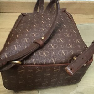 K&ouml;nnte beinhalten: Ein brauner Lederrucksack mit einem Monogrammbild. Der Rucksack hat verstellbare Tr&auml;ger und einen oberen Griff.