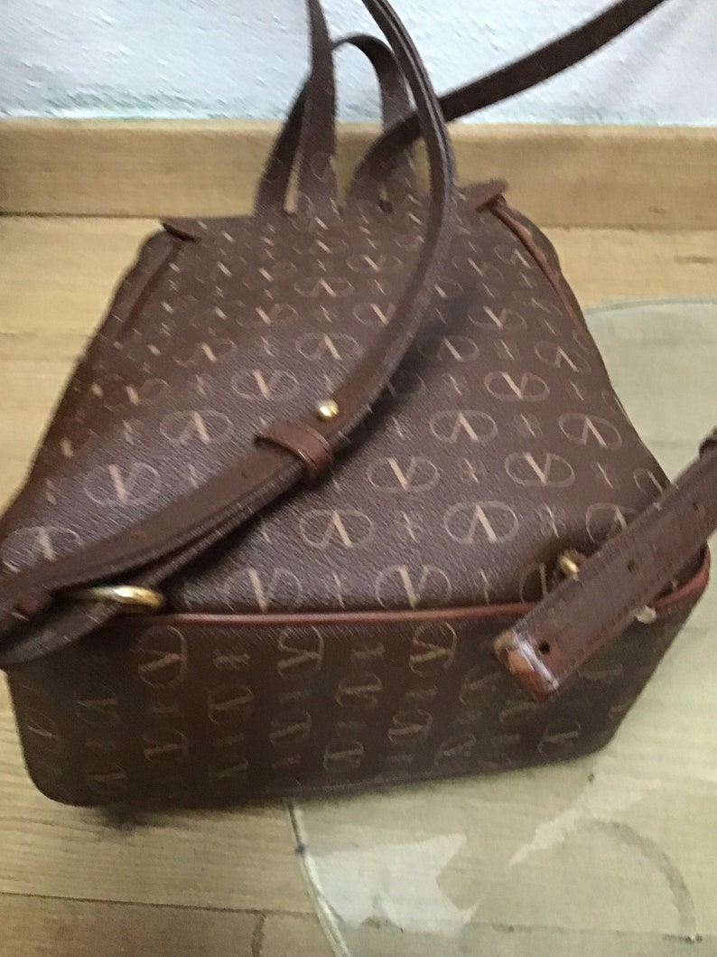 K&ouml;nnte beinhalten: Ein brauner Lederrucksack mit einem Monogrammbild. Der Rucksack hat verstellbare Tr&auml;ger und einen oberen Griff.