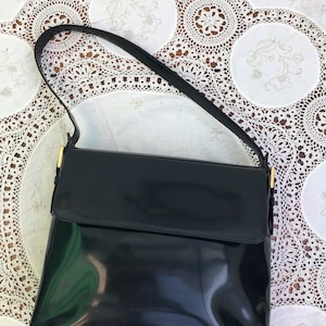Puede incluir: Un bolso de mano de charol negro con cierre de solapa rectangular y una sola correa para el hombro. El bolso cuenta con herrajes de metal en tono dorado que conectan la correa con el cuerpo principal, ofreciendo un acabado brillante.