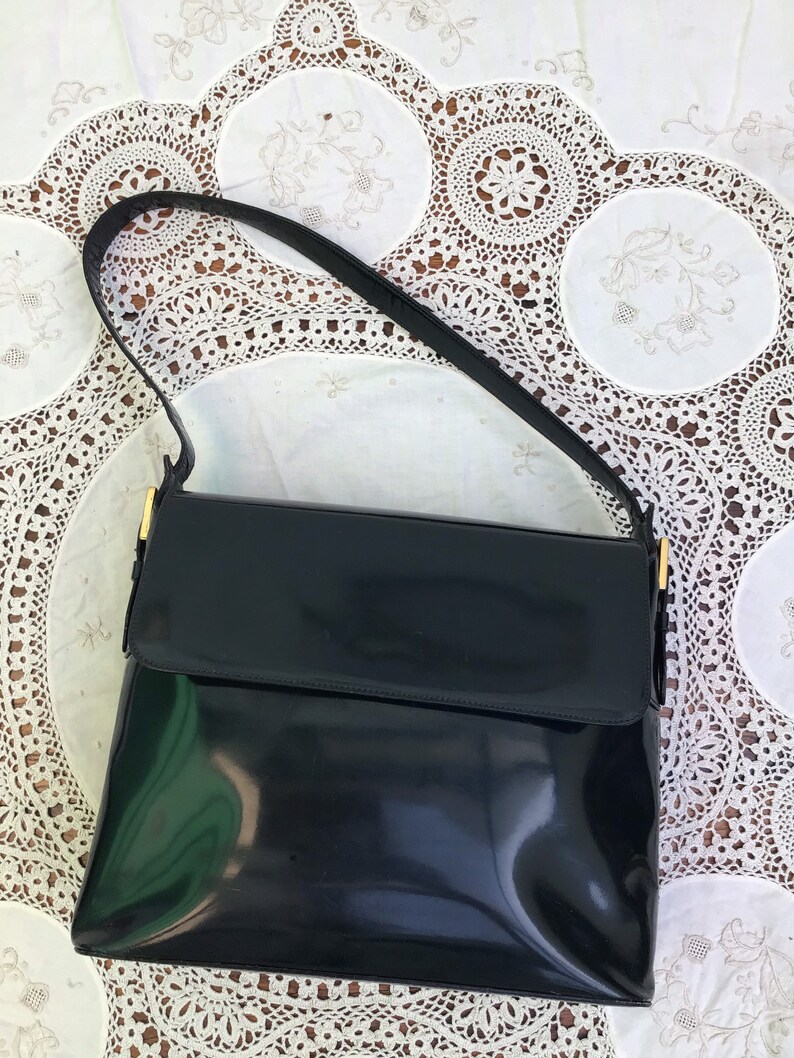 Puede incluir: Un bolso de mano de charol negro con cierre de solapa rectangular y una sola correa para el hombro. El bolso cuenta con herrajes de metal en tono dorado que conectan la correa con el cuerpo principal, ofreciendo un acabado brillante.