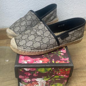 Può includere: Un paio di scarpe slip-on in stile espadrillas con un motivo a logo ripetuto nei toni del marrone e beige. Le scarpe hanno un bordo nero e una suola in juta intrecciata. Sono appoggiate su una scatola a motivi floreali. Misurano circa 25 cm.