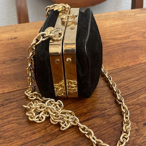 Op de afbeelding: Zwarte suède clutch met goudkleurige metalen accenten en een kettingriem. De rechthoekige tas heeft een afgeronde vorm en een sluiting met gesp. De gouden ketting is dik en voegt elegantie toe aan het ontwerp.