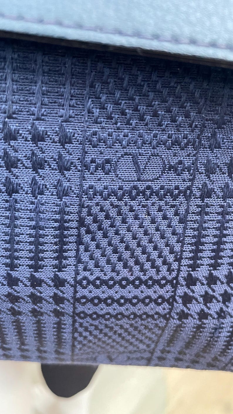 May include: Close-up of a textured, navy blue fabric with a repeating geometric pattern. The fabric appears to be woven, with a subtle sheen. The design is intricate and detailed, creating a visually interesting surface.