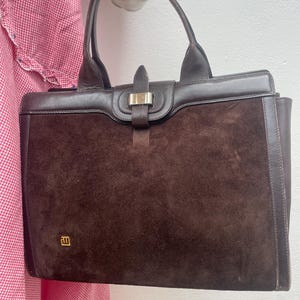 Può includere: Una borsa in camoscio marrone con forma rettangolare e manico superiore. La borsa presenta finiture in pelle marrone scuro e una chiusura color oro. Un piccolo emblema dorato è visibile sulla parte anteriore inferiore della borsa. La borsa è tenuta da una mano decorativa.