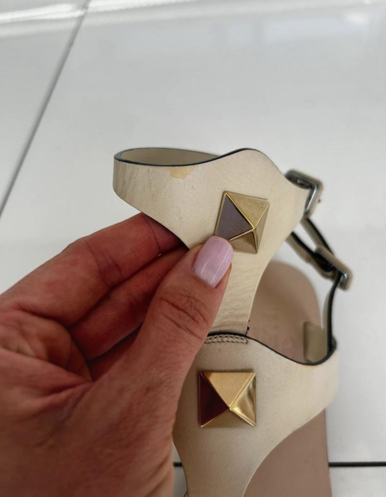 May include: Cream-colored leather sandals with gold pyramid studs. The sandals have a T-strap design and a buckle closure. The studs are a prominent feature, adding a touch of luxury to the footwear. The sandals appear to be new and unworn.