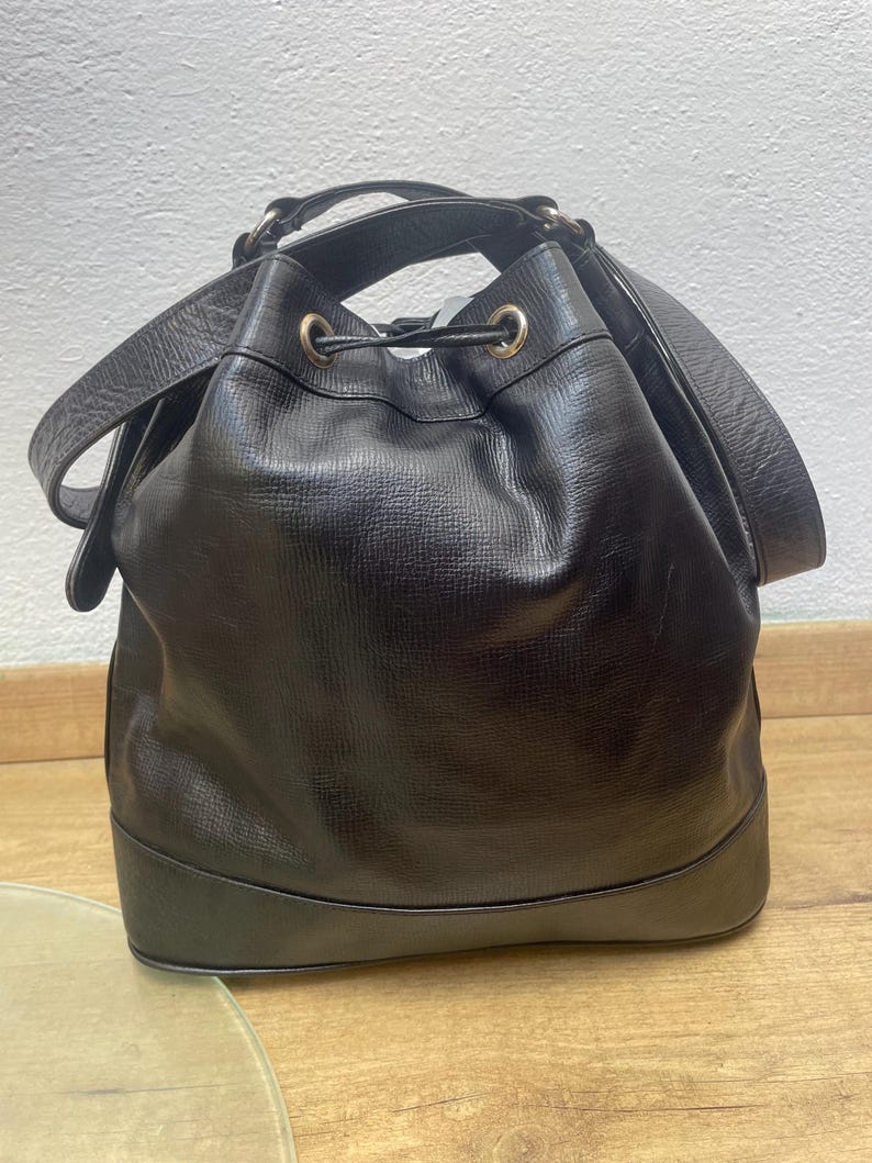 May include: Black leather bucket bag with a drawstring closure and shoulder strap. The bag has a rounded base and gold-toned hardware. The leather has a textured appearance.
