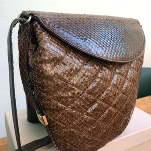 Op de afbeelding: Bruine, getextureerde crossbodytas met een klepsluiting. De tas heeft een slangenpatroon en een lange, verstelbare riem. Een goudkleurige sluiting zekert de klep. De tas staat op een wit oppervlak.