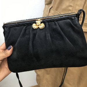 Può includere: Una pochette in camoscio nero con chiusura dorata e un gruppo decorativo di tre abbellimenti dorati tempestati di strass. La pochette ha una lunga e sottile tracolla nera e un bordo superiore strutturato.