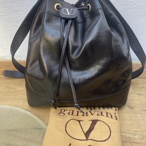 May include: A black leather bucket bag with a drawstring closure and shoulder straps. The bag features gold-tone hardware and a silver logo. A tan dust bag with the brand's logo is visible.