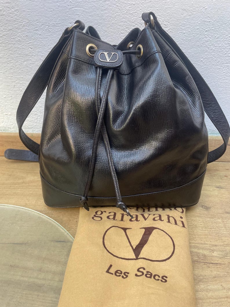 May include: A black leather bucket bag with a drawstring closure and shoulder straps. The bag features gold-tone hardware and a silver logo. A tan dust bag with the brand's logo is visible.