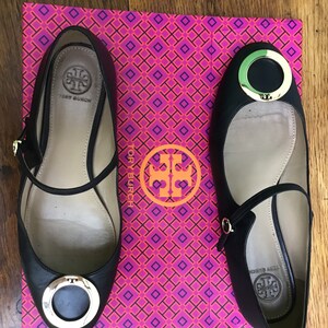 May include: Black leather ballet flats with gold-tone hardware, featuring a round toe and ankle strap. The shoes are displayed on a pink and orange patterned box with the brand logo.