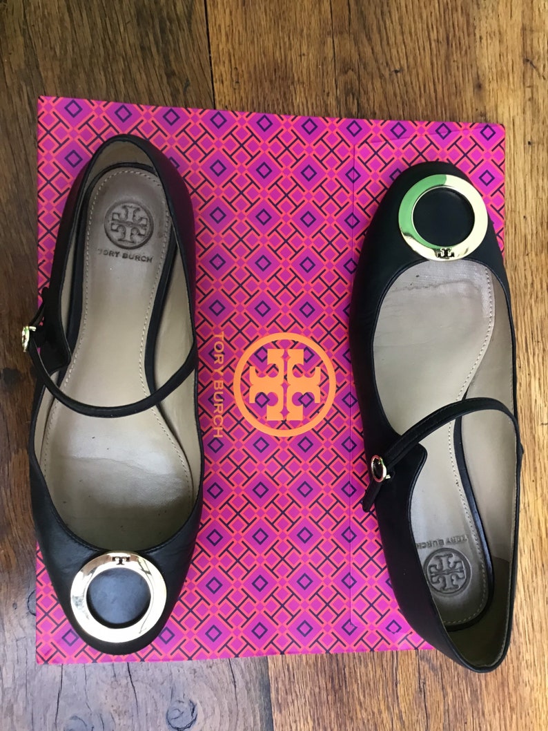 May include: Black leather ballet flats with gold-tone hardware, featuring a round toe and ankle strap. The shoes are displayed on a pink and orange patterned box with the brand logo.