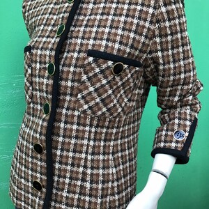 Puede incluir: Una chaqueta de cuadros marrones, negros y blancos con cuello redondo, mangas largas y cierre frontal de botones. La prenda incluye dos bolsillos en el pecho con ribete negro, bordes con ribetes negros y botones negros con borde dorado.