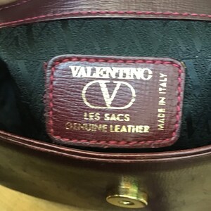 Może przedstawiać: Zbliżenie na wnętrze bordowej sk&oacute;rzanej torebki. Etykieta głosi "VALENTINO LES SACS GENUINE LEATHER MADE IN ITALY" złotymi literami. Torba ma czarne wnętrze i złote zapięcie.