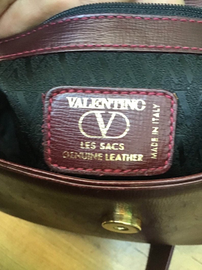 Może przedstawiać: Zbliżenie na wnętrze bordowej sk&oacute;rzanej torebki. Etykieta głosi "VALENTINO LES SACS GENUINE LEATHER MADE IN ITALY" złotymi literami. Torba ma czarne wnętrze i złote zapięcie.