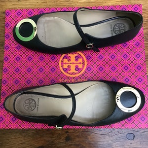 May include: Black leather ballet flats with a gold-tone circular accent and ankle strap. The shoes are displayed on a pink and orange patterned box with a logo. The flats have a rounded toe and a small heel.