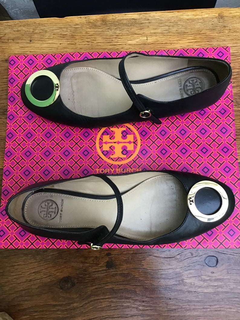 May include: Black leather ballet flats with a gold-tone circular accent and ankle strap. The shoes are displayed on a pink and orange patterned box with a logo. The flats have a rounded toe and a small heel.