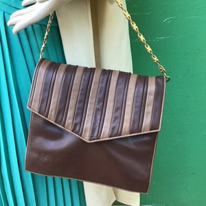 May include: A brown leather handbag with a flap closure and a gold chain strap. The top of the bag features vertical stripes in shades of brown and tan. The bag has a logo on the lower front.