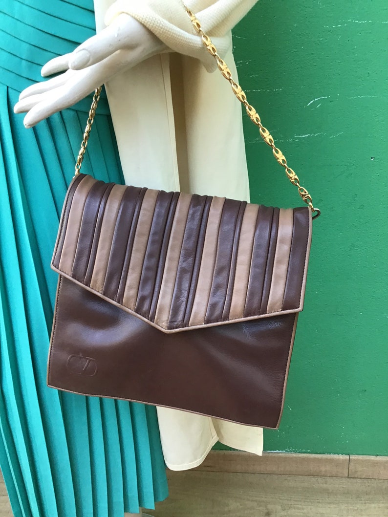 May include: A brown leather handbag with a flap closure and a gold chain strap. The top of the bag features vertical stripes in shades of brown and tan. The bag has a logo on the lower front.